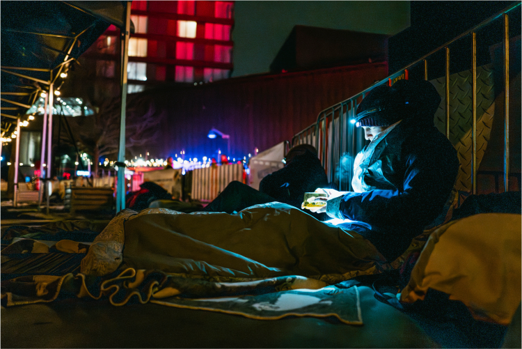 Een deelnemer aan de Sleep Out ligt in Sheltersuit en muts tegen een hek (C) Sheltersuit Foundation